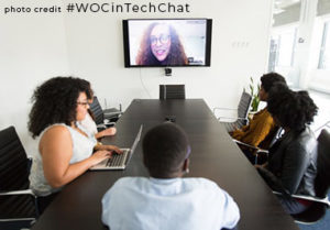Black women sitting at a boardroom table looking at a television screen for a videoconference.